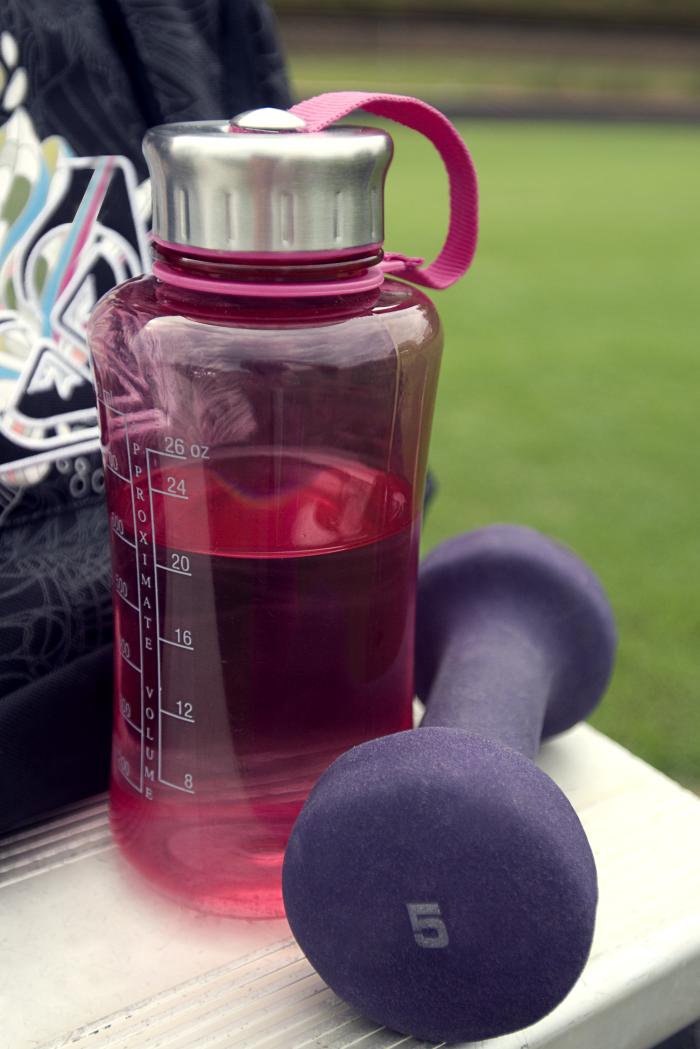 Taken at an outdoor Georgia running track, this image depicts a partially-filled red plastic water bottle, along-side a 5lb, purple colored dumbbell.