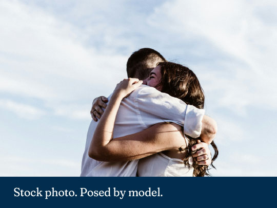 Photograph of two people embracing outdoors against a partly cloudy sky, conveying affection. Both individuals wear light-colored clothing, with one person's face visible showing a joyful expression. Text 'Stock photo. Posed by model.'