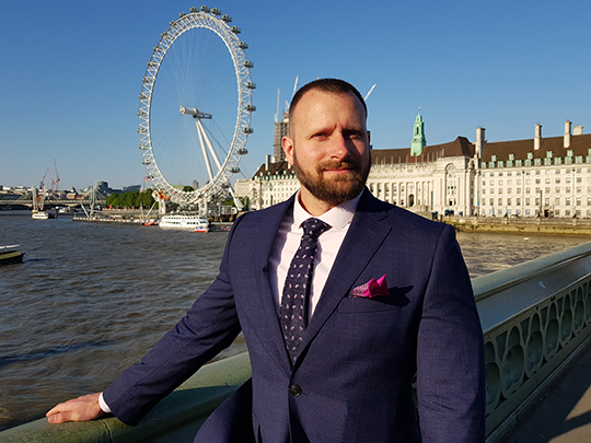 Photo of David Vaughan: a man in a suit posing in front of a bridge.